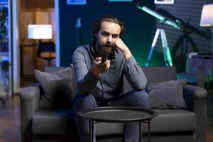 cansado homem zapping através canais, assistindo notícia transmissão enquanto ocioso dentro vivo quarto depois de Difícil dia às trabalhar. pessoa usando controlo remoto ao controle para selecionar jornalismo televisão estação para apreciar foto