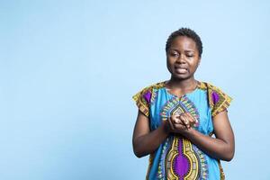 africano americano mulher Rezar e falando para Jesus dentro estúdio, segurando mãos dentro uma oração enquanto ela desgasta tradicional étnico roupas. jovem fêmea modelo ser espiritual e religioso em Câmera. foto