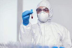 homem dentro macacão terno dentro microbiologia laboratório segurando teste tubo com sangue infectado com coronavírus. médico trabalhando com vários bactérias e tecido, farmacêutico pesquisa para antibióticos contra COVID-19. foto