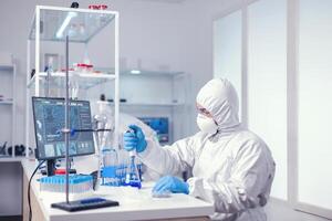 com experiência biólogo vestido dentro ppe segurando pipeta com azul solução. químico dentro moderno laboratório fazendo pesquisa usando distribuidor durante global epidemia com COVID-19. foto