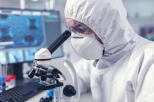 fechar acima do investigador olhando através microscópio dentro bioquímica laboratório. virólogo dentro macacão durante coronavírus surto conduzindo cuidados de saúde científico análise. foto