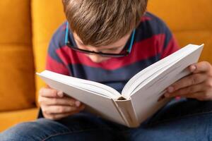 jovem estudante com óculos imerso dentro livro lendo em sofá foto