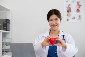 feliz jovem médico Senior mulher segurando vermelho coração forma objeto, olhando às Câmera com sorriso. positivo praticante, cardiologista foto