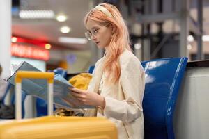 feliz jovem mulher ásia é sentado dentro aeroporto perto mala de viagem e lendo mapa foto