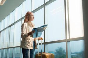 feliz jovem mulher ásia é ficar de pé dentro aeroporto perto mala de viagem e lendo mapa foto