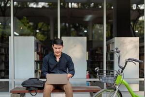 ásia homem de negocios com bicicleta usando computador portátil e sentado lado de fora a escritório prédio. homem pendulares em bicicleta ir para trabalhar. eco amigáveis veículo, sustentável estilo de vida conceito foto