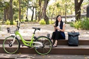 ásia empresária com bicicleta usando Smartphone e sentado lado de fora a escritório prédio. mulher pendulares em bicicleta ir para trabalhar. eco amigáveis veículo, sustentável estilo de vida conceito foto