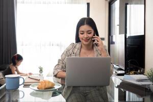 ásia fêmea Falando em Móvel telefone enquanto usando computador portátil para controlo remoto trabalhos levando Cuidado do filha às casa foto