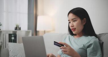 seletivo foco, jovem ásia mulher sentado em sofá segurando crédito cartão fazer conectados Forma de pagamento para compra dentro rede loja usando computador portátil. foto