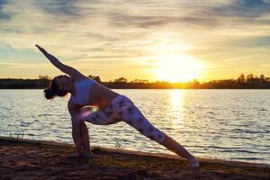 jovem mulher fazendo ioga exercícios em a lago de praia às pôr do sol foto