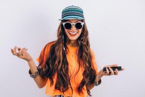 jovem hipster lindo mulher dentro oculos de sol vestindo dentro boné e laranja camiseta ouvindo música dentro fones de ouvido perto branco parede, segurando uma célula telefone dentro mão foto