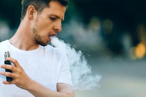 moderno jovem homem com uma barba Diversão vaporizadores ar livre. masculino dentro branco camiseta vape ecig. Sombrio fundo, a tarde pôr do sol sobre a cidade. tonificado imagem. foto