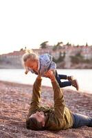 Papai é deitado em a de praia segurando uma pequeno menina acima ele dentro estendido braços foto
