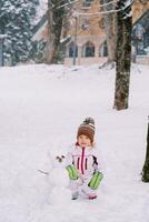 pequeno menina agachamentos perto uma pequeno boneco de neve dentro uma Nevado floresta foto