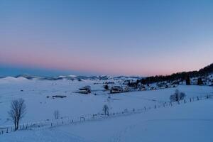 rosa-azul pôr do sol sobre uma coberto de neve Vila em a Beira do uma floresta dentro uma montanha vale foto