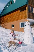 pequeno menina come mingau a partir de uma placa, sentado em uma trenó perto uma boneco de neve perto a casa foto