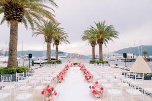 linhas do cadeiras linha uma caminho decorado com vermelho flores dentro frente do uma Casamento arco em a cais foto