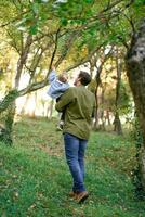 pequeno menina dentro dela pai braços pontos dela dedo às uma árvore dentro a parque. costas Visão foto
