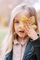 pequeno menina cobertura metade do dela face com uma amarelo folha enquanto em pé dentro a outono parque foto