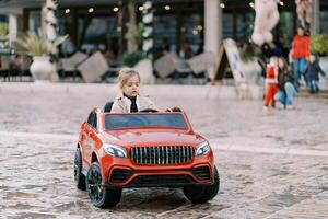 pequeno menina drives uma vermelho brinquedo carro através a quadrado foto