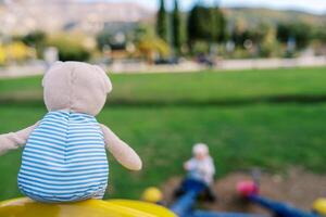 Urso de pelúcia Urso dentro uma listrado camiseta senta em uma balanço dentro a Parque infantil. costas Visão foto