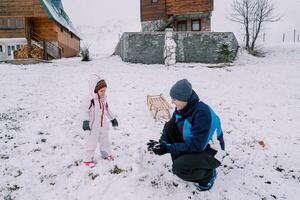 pequeno menina parece às dela Papai fazer uma bola de neve em dele ancas perto uma de madeira casa foto