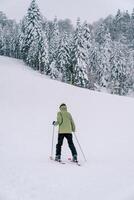 esquiador dentro uma esqui terno esquis acima uma coberto de neve montanha ao longo a floresta. costas Visão foto