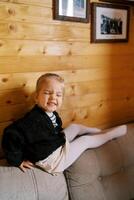 pequeno sorridente menina sentado em a costas do uma sofá inclinado contra uma de madeira parede foto