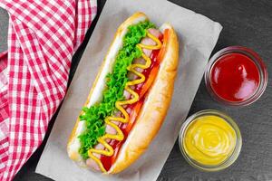 caseiro quente cachorro com mostarda, ketchup, tomate e fresco salada folhas em Preto ardósia fundo foto