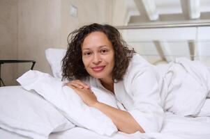 jovem mulher deitado dentro branco cama em branco travesseiro e coberto com confortável suave cama folhas, sorridente olhando às Câmera foto
