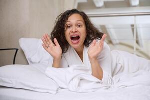 retrato do a animado jovem mulher deitado em a cama dentro a manhã, desfrutando dela final de semana às casa foto