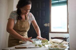 lindo dona de casa usando vidro vinho garrafa Como rolando alfinete, rolando Fora verde espinafre massa para fazer dumplings, em pé às cozinha mesa borrifado com farinha dentro a rural casa cozinha foto