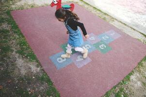 a sobrecarga Visão criança menina jogando amarelinha em a escola Parque infantil. popular rua crianças jogos dentro clássicos foto