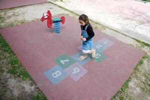 Visão a partir de acima do uma criança menina jogando amarelinha, levando voltas pulando sobre a quadrados marcado em a escola jogar chão. foto