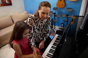 Visão a partir de acima pequeno menina sentado às grande piano perto dela professor, tendo piano lição às lar. diverso musical instrumentos contra uma azul cor parede em a fundo. pessoas. Educação. estilo de vida foto