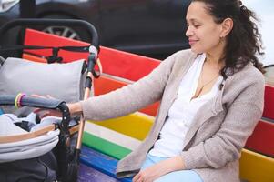 jovem mãe sorridente, empurrando bebê carrinho de bebê com dela dormindo bebê, sentado em lgbt banco. tolerância, igualdade, liberdade foto