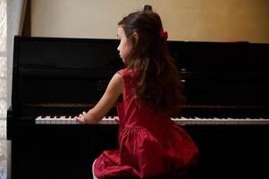 autêntico caucasiano pequeno criança menina músico pianista com lindo grandes cabelo, vestido dentro elegante vermelho vestir, colocando dedos em piano chaves, desfrutando a desempenho do clássico música. cópia de de Anúncios espaço foto