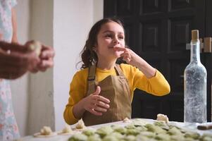 caucasiano pernicioso pequeno criança menina dentro bege avental, sorridente enquanto degustação amassado batata enquanto cozinhando dumplings com dela mãe dentro a casa cozinha. foto