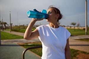 desportivo Atlético mulher bebendo água a partir de Esportes garrafa enquanto relaxante depois de ao ar livre exercite-se em caloroso ensolarado dia foto