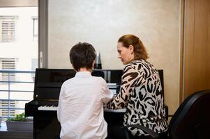 traseiro Visão do uma música professor explicando piano lição para dela estudante. Garoto jogando piano debaixo a orientação do hos professor durante Individual música lição às casa foto