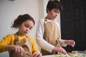 fofa crianças Garoto e menina dentro a rural cozinha, escultura dumplings a partir de massa com amassado batatas enchimento. cozinhando caseiro vegetariano dumplings, italiano Ravioli ou ucraniano Varenyky foto