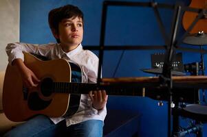 hispânico Adolescência Garoto dentro branco casual camisa, arrancar cordas enquanto dedilhar guitarra, jogando acústico clássico guitarra, sentado dentro a música estúdio sobre azul parede fundo e musical instrumentos foto