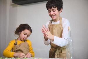 fofa crianças dentro bege chefe de cozinha avental, fazer dumplings durante uma cozinhando classe dentro de casa, em pé contra uma branco parede fundo foto