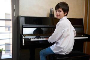 feliz adolescente Garoto dentro branco casual camisa, sentado de piano e sorridente às Câmera enquanto realizando melodia em a piano forte foto