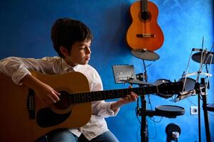 adolescente Garoto dedilhar guitarra dentro a moderno música estúdio. elétrico guitarra suspensão em uma azul parede perto bateria. pessoas. música lição. crianças Educação e entretenimento foto