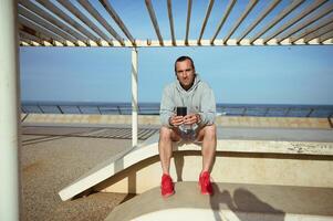jovem homem dentro Esportes roupas, usando Móvel telefone, sentado em pedra Banco depois de exercite-se foto