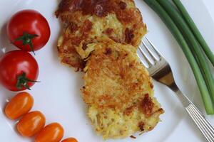 fechar-se do delicioso caseiro frito Grato batata panquecas servido com fresco tomates e verde cebolas em a branco placa, garfo em a certo lado. draniki. vegetal refeição. topo visualizar. foto