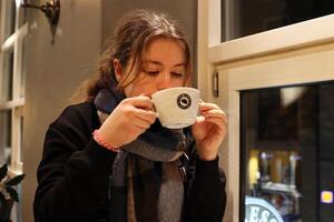 copenhague, Dinamarca - fevereiro, 20, 2023. espresso casa. cabeludo adolescente menina é bebendo uma copo do café sentado Próximo para janela. tarde rua luzes e acolhedor atmosfera do a café fazer compras. foto