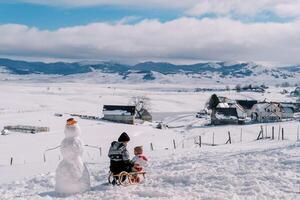 mãe e pequeno menina estão sentado em uma trenó em uma coberto de neve avião perto uma boneco de neve e Veja às cada outro. costas Visão foto