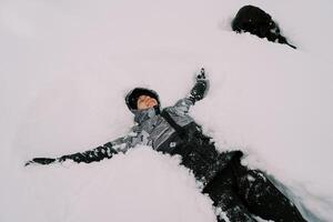 jovem sorridente mulher dentro uma esqui terno mentiras dentro a neve com dela braços estendido perto uma Preto cachorro foto